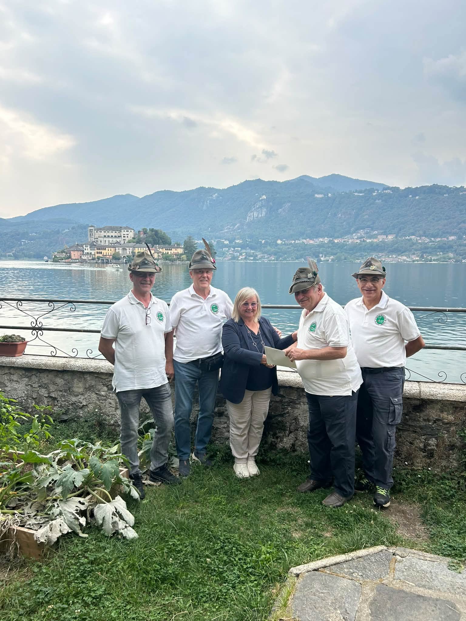 Quattro alpini con il cappello e una signora al centro che tiene un foglio, in piedi davanti a un muretto con ringhiera; dietro, il lago con un’isola e le colline