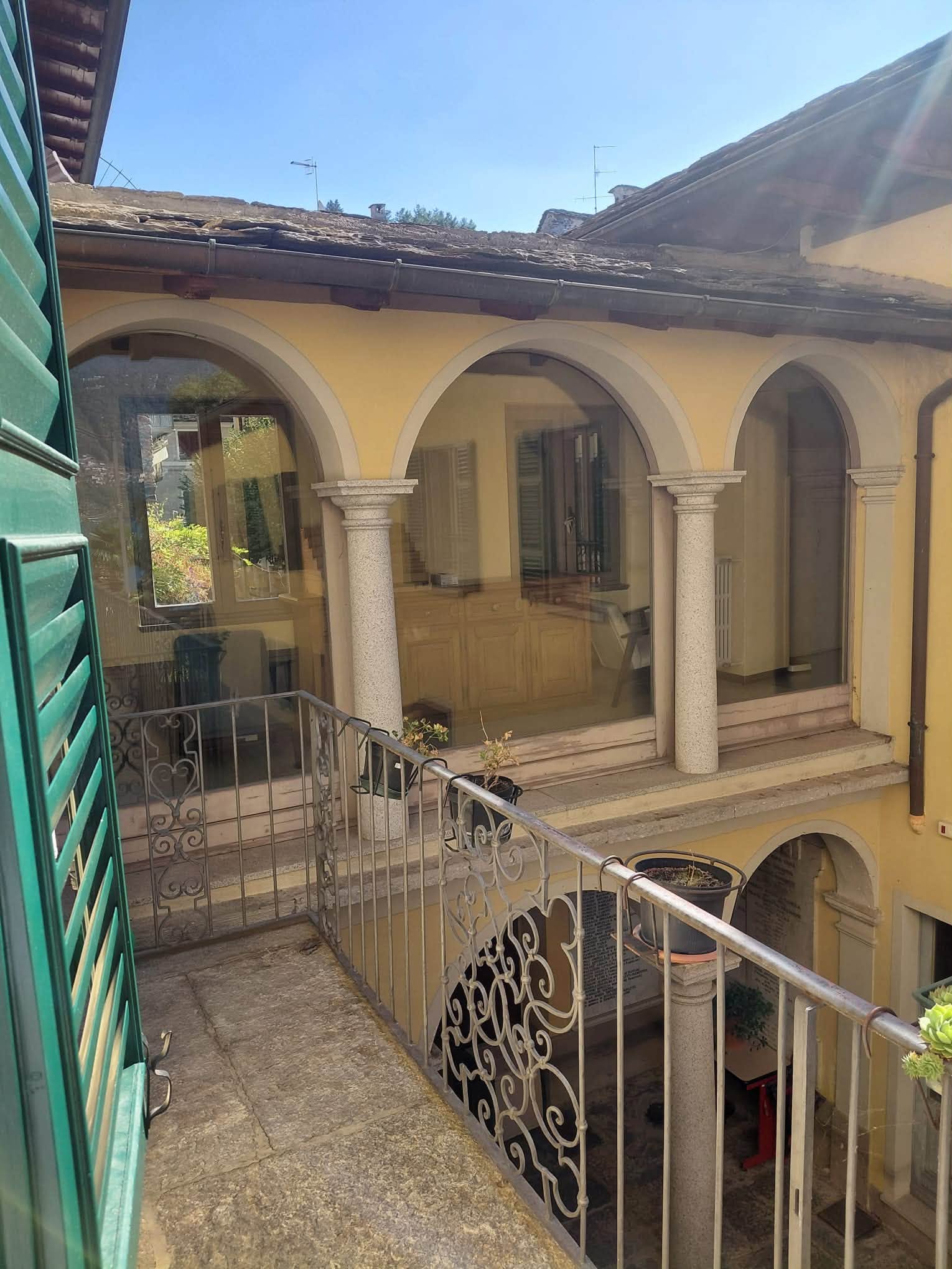 La corte interna vista da un balcone con le persiane verdi: l’edificio giallo con il portico ad archi e la ringhiera in ferro battuto