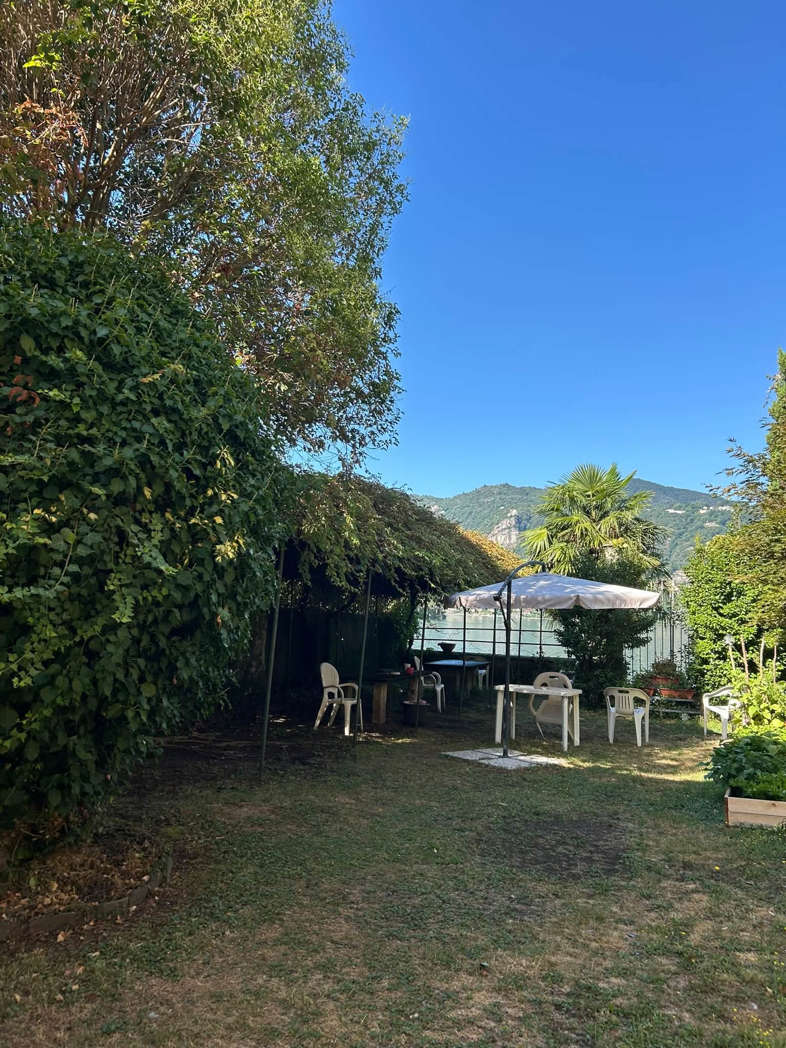 Il giardino: tavoli e sedie sotto un ombrellone e un pergolato, con il lago e i monti sullo sfondo